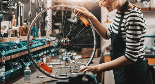Fototapeta Naklejka Na Ścianę i Meble -  Cute Caucasian female worker holding and repairing bicycle wheel while standing in bicycle workshop.