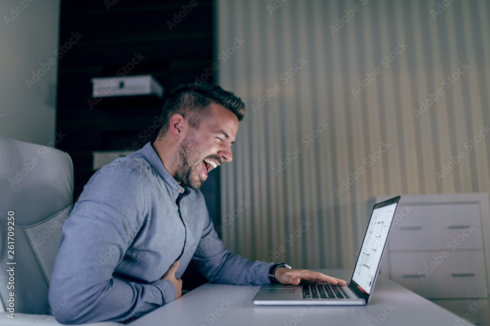 Laughing handsome Caucasian bearded architect sitting in office at night holding hand on laptop. Laughing handsome Caucasian bearded architect sitting in office at night holding hand on laptop.