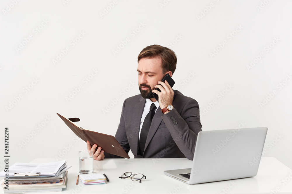 Busy businessman, top manager with beard attentively looking through notes in his diary sitting at desktop in office, arrange meeting with mobile phone, dressed in a suit with a tie, has a good watch Busy businessman, top manager with beard attentively looking through notes in his diary sitting at desktop in office, arrange meeting with mobile phone, dressed in a suit with a tie, has a good watch