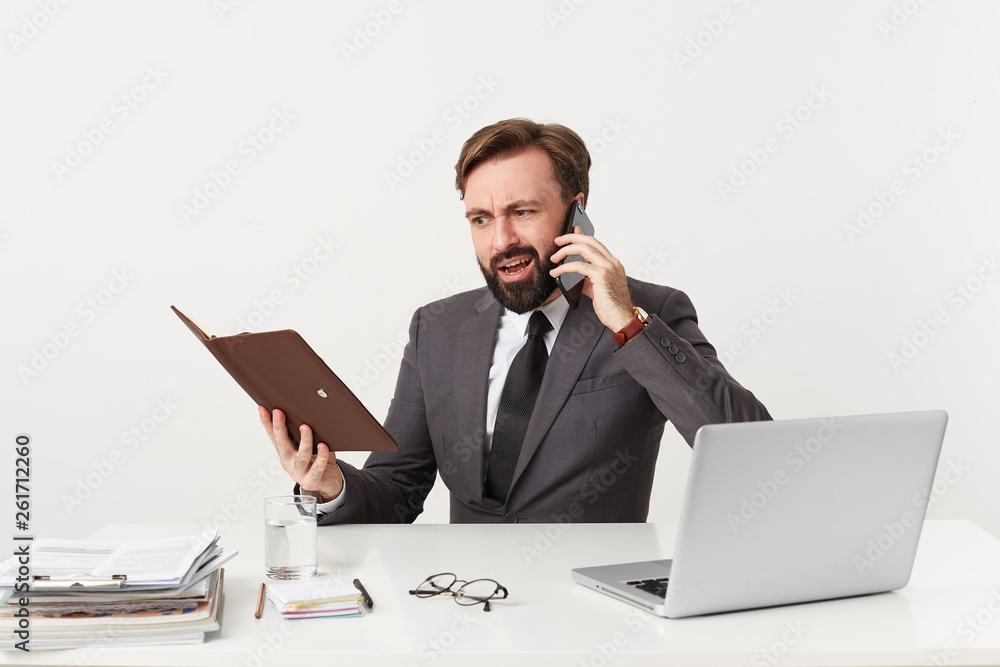 Businessman, top manager with beard looking through notes in his diary sitting at desktop in office, has unpleasant telephone conversation, argues quarrel with someone, dressed in a suit with a tie Businessman, top manager with beard looking through notes in his diary sitting at desktop in office, has unpleasant telephone conversation, argues quarrel with someone, dressed in a suit with a tie