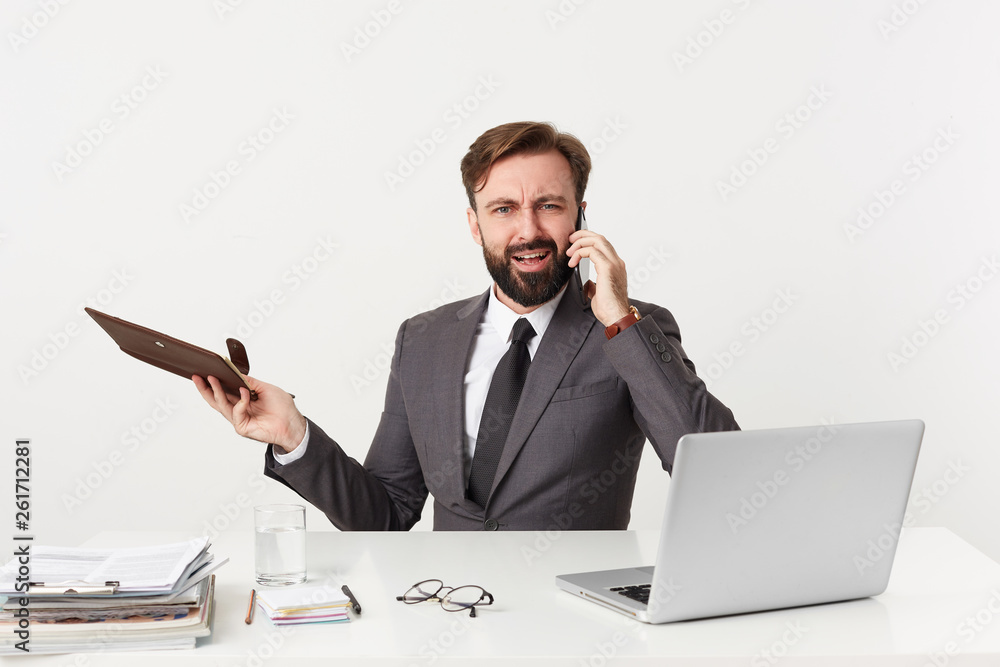 Businessman, top manager with beard keeps his notebook sitting at table in office, looking camera while has unpleasant telephone conversation, argues quarrel with someone, dressed in a suit with a tie Businessman, top manager with beard keeps his notebook sitting at table in office, looking camera while has unpleasant telephone conversation, argues quarrel with someone, dressed in a suit with a tie