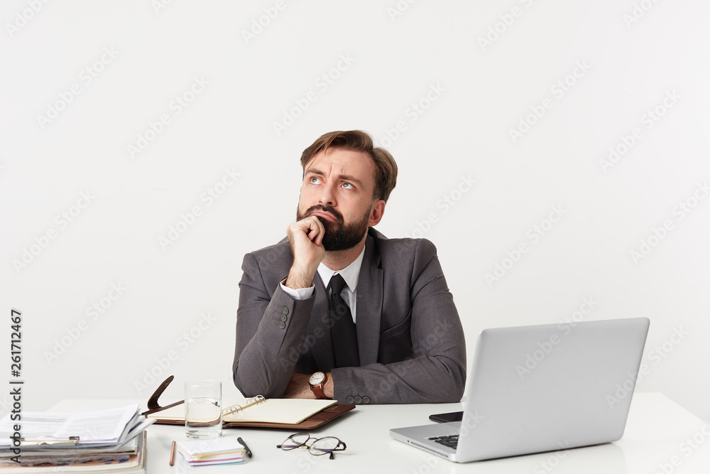 Thoughtful manager with beard sits at the table props his chin with his hand looks to the left side bored doesn't know what to do, ponders about something unimportant, dressed in a suit with a tie Thoughtful manager with beard sits at the table props his chin with his hand looks to the left side bored doesn't know what to do, ponders about something unimportant, dressed in a suit with a tie