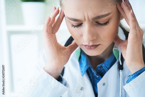 Woman doctor with migraine headache, overworked and stressed. Health care professional in lab coat wearing stethoscope at hospital. © cameravit