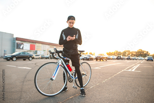 Young man is in a dark sporty attire with a smartphone in the hands of a parking lot and looks at the screen. Happy young cyclist uses a smartphone on the background and the sunset.