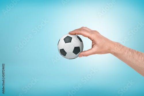 Male hand holding small football ball between fingers on blue background