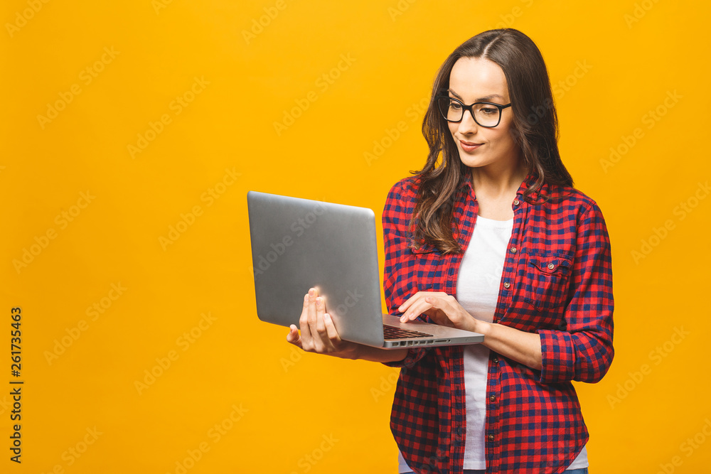 Naklejka premium Young happy smiling woman in casual clothes holding laptop and sending email to her best friend. Isolated against yellow background.