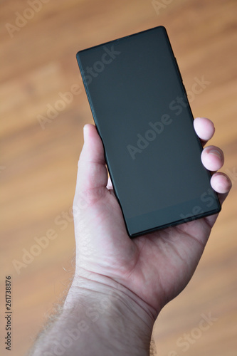 The male hand holds the smartphone with the dark screen against the background of an indistinct wooden surface