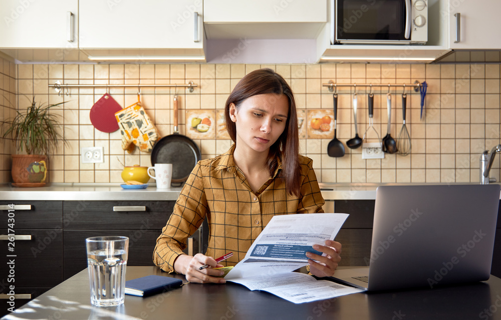 Thoughtful stressed young female sitting at kitchen table with papers and laptop computer trying to work through pile of bills, frustrated by amount of domestic expenses while doing family budget 