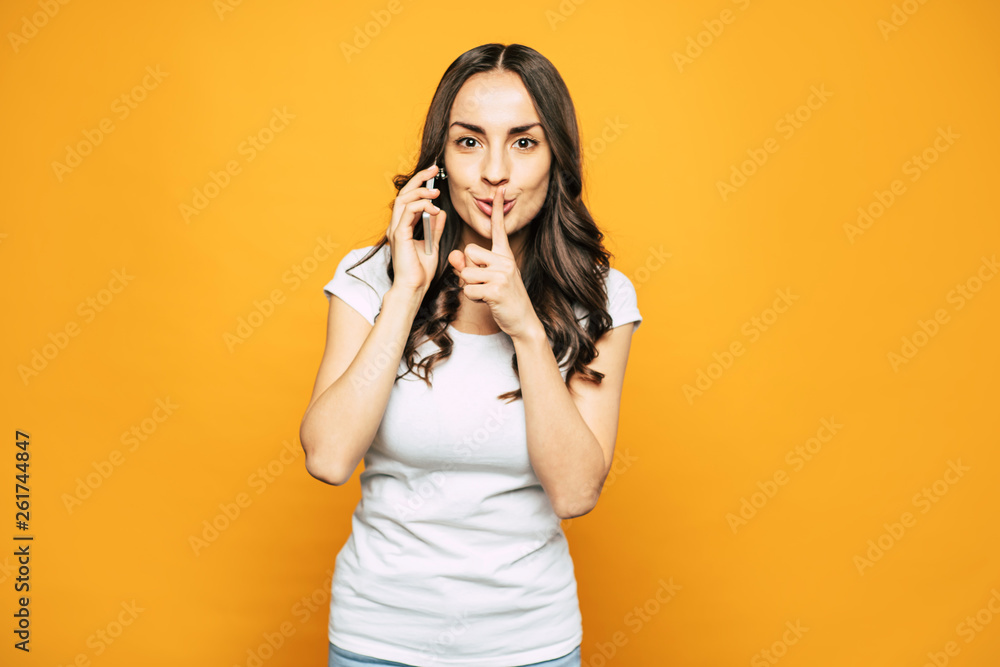 SHHH! Nice looking girl with glowing skin, slightly curled hair and elegant smile next to the flame-colored background is showing “shh” because she is having a phone call. SHHH! Nice looking girl with glowing skin, slightly curled hair and elegant smile next to the flame-colored background is showing “shh” because she is having a phone call.