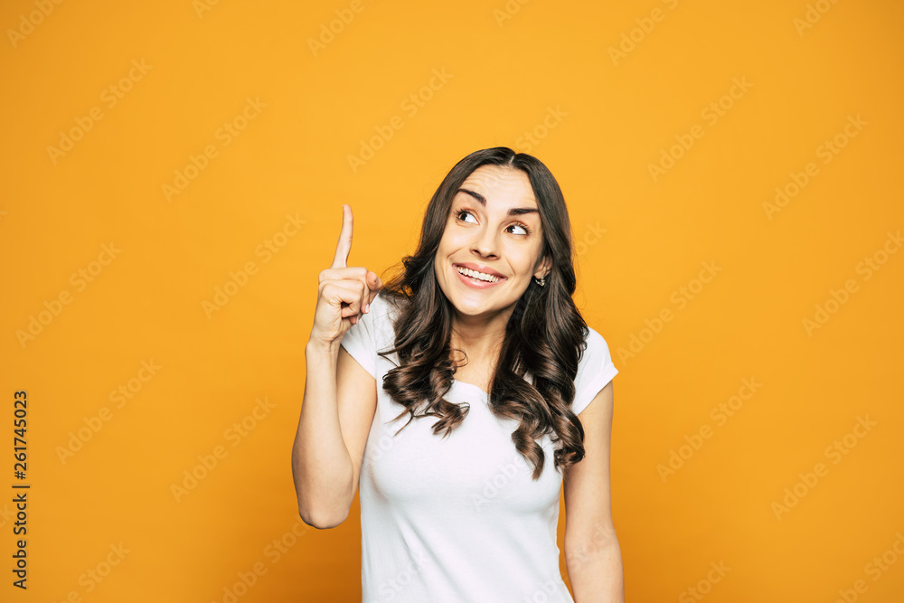 Obraz premium Idea! Pretty girl with nice hairstyle and gorgeous smile in white T-shirt is holding her finger pointed up which is telling about her genius idea.