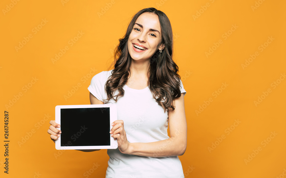 Here it is! A girl with sincere brown eyes, dark-brown wavy long hair near the vibrant orange background is holding a tablet PC in her elegant hands. Here it is! A girl with sincere brown eyes, dark-brown wavy long hair near the vibrant orange background is holding a tablet PC in her elegant hands.