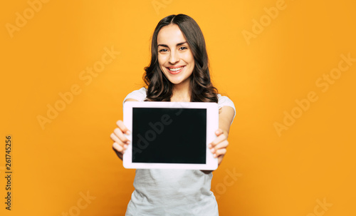 Come and see. A tablet PC in the hands of bodacious smiley girl with slightly curled brownish hair, hazel eyes and stunning smile in front of spicy-orange background. photo