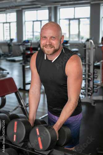 handsome young man in blue shorts and black t-shirts, doing exercises for biceps in the gym. bald man smiling at camera