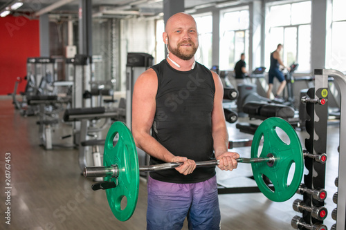 Bald athlete in a black T-shirt and blue shorts with headphones, lifting the barbell with effort. He is standing in the gym and smiling