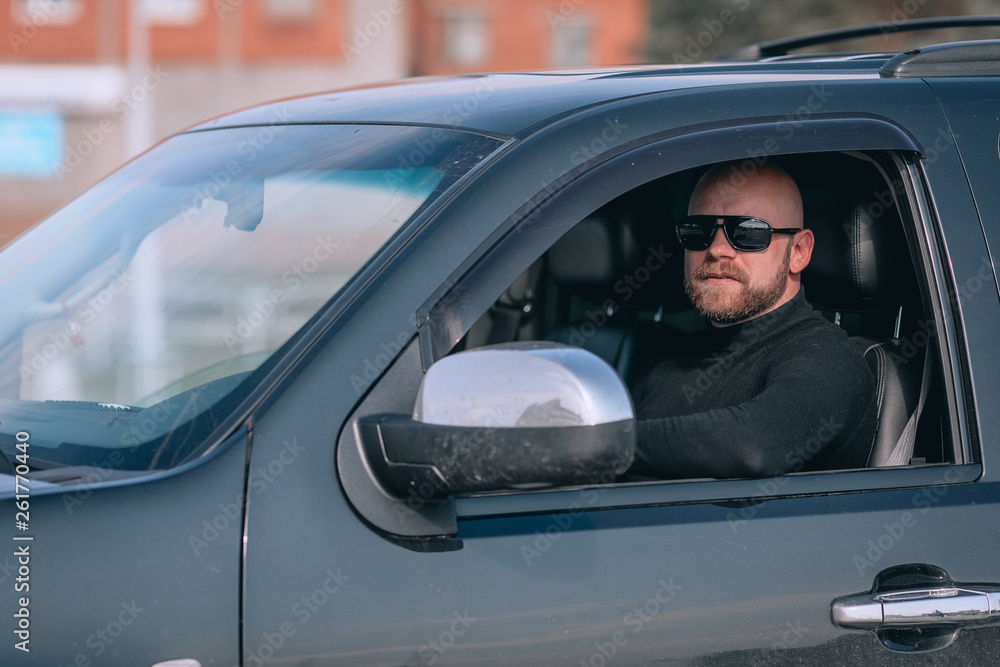 Bald and bearded man in glasses with a clock in a suit behind the wheel of a black car. motorists concept Bald and bearded man in glasses with a clock in a suit behind the wheel of a black car. motorists concept
