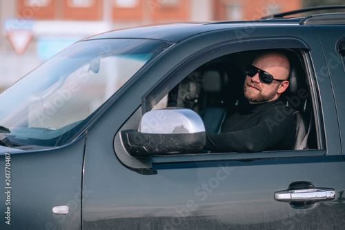 bald and bearded human in glasses with a clock in a suit behind the wheel of a black car