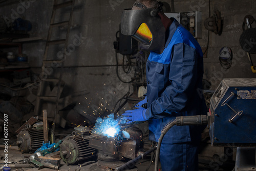 adult welder is welding a metal construction in garage wearing mask, proctive glasses and blue uniform. Blue sparks are flying apart. © Semachkovsky 