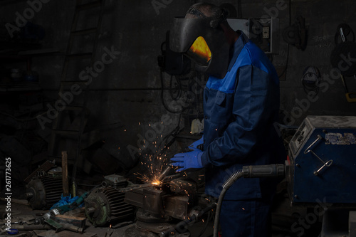  welder is welding a metal construction in garage wearing mask, proctive glasses and blue uniform. Blue sparks are flying apart.
