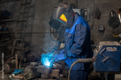 Strong man is welding a metal construction in garage wearing mask, proctive glasses and blue uniform. Blue sparks are flying apart.