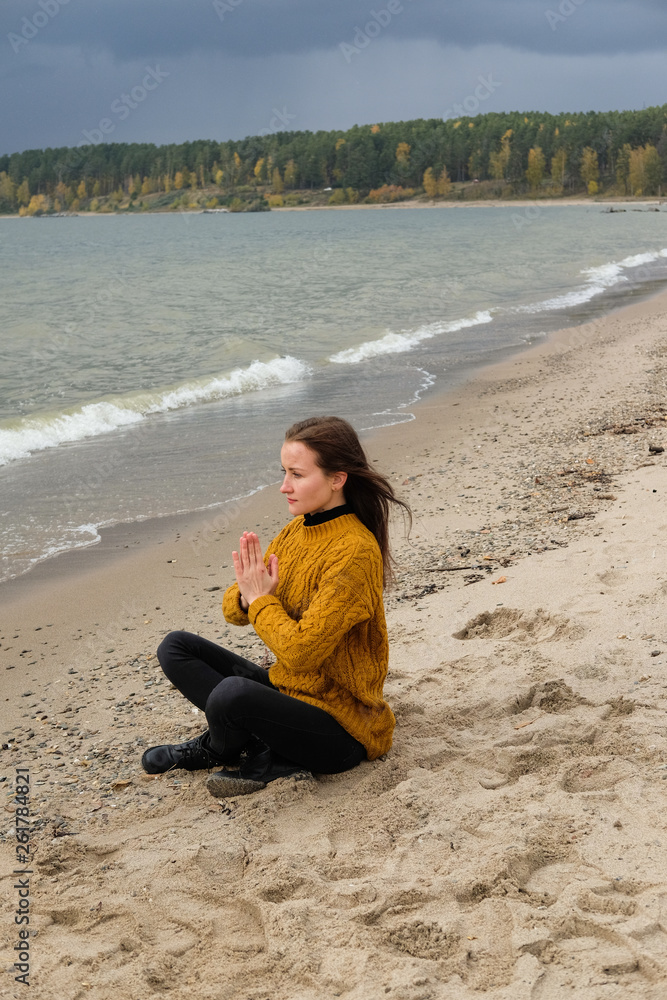 Woman practicing yoga against the blue sky and the azure sea on the sandy shore. Woman raises her arms to the sky in namaste posture and sits in a lotus pose.