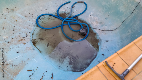 Bottom of drained swimming pool with just a little dirty water around the drain and a vacuum hose curled nearby with section of tile deck and cleaning brush visible