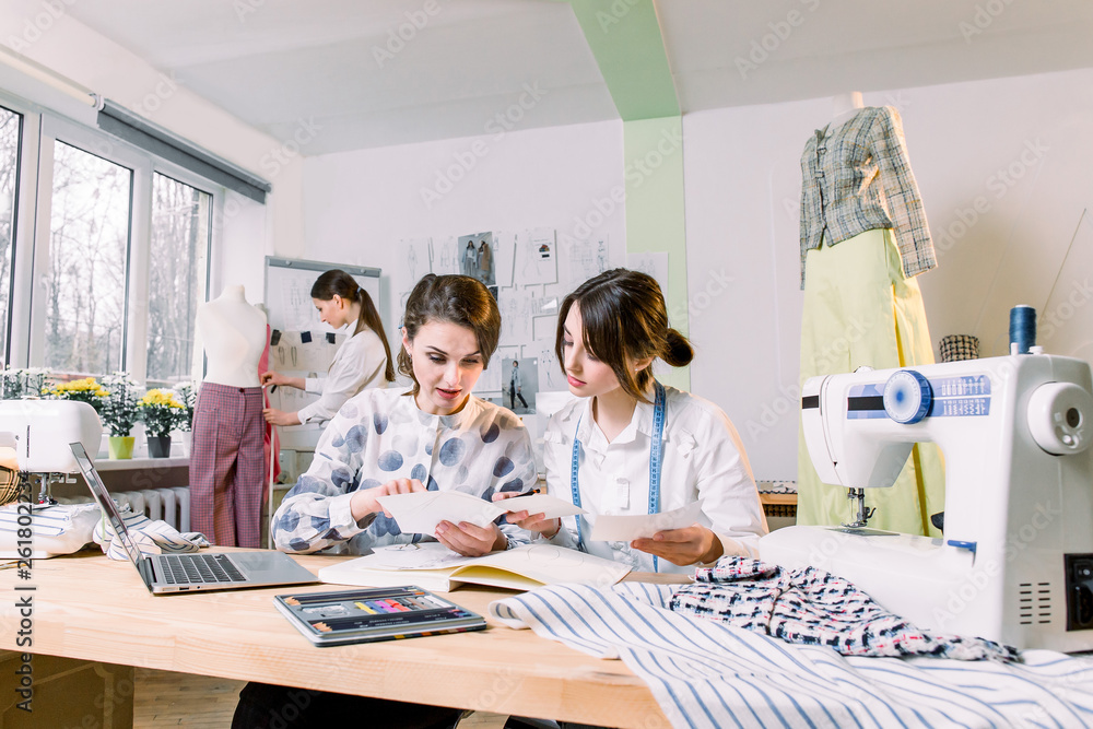 Two attractive women dressmakers designers choosing trendy sketches and materials for new collection of clothes, while young girl designer working on new model tailoring pants on mannequin in studio Two attractive women dressmakers designers choosing trendy sketches and materials for new collection of clothes, while young girl designer working on new model tailoring pants on mannequin in studio