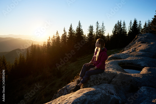 Young woman traveller sitting on big boulder on the top of mountain in the evening. Female tourist enjoying beautiful sunset. On background forest, setting sun and blue sky