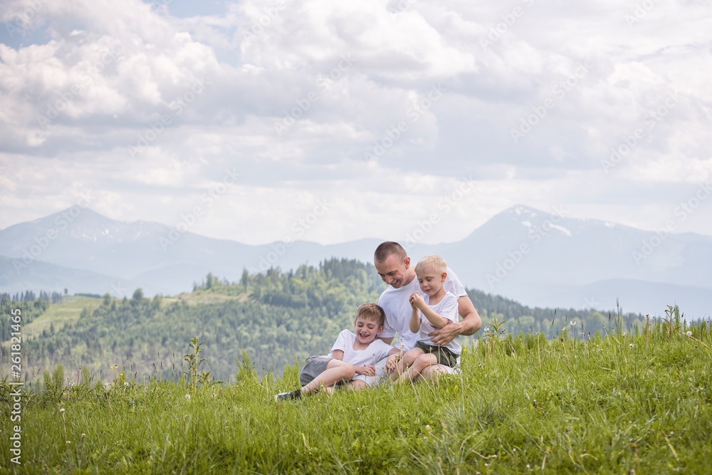 Happy father with his two young sons sitting on the grass on a background of green forest, mountains and sky with clouds. Friendship concept. Happy father with his two young sons sitting on the grass on a background of green forest, mountains and sky with clouds. Friendship concept.