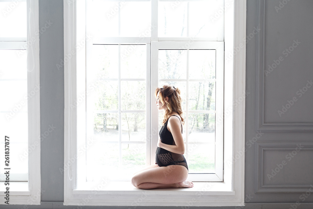 Side view portrait of beautiful pregnant young woman in black bodysuit sitting in front of window at home and holding with love her belly. Last months of pregnancy. Studio shot, light background Side view portrait of beautiful pregnant young woman in black bodysuit sitting in front of window at home and holding with love her belly. Last months of pregnancy. Studio shot, light background