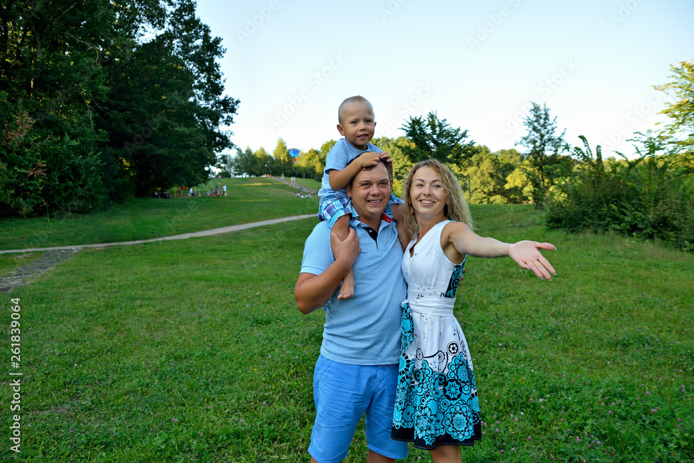 Naklejka premium Happy family with a child walking in the Park outdoors. A little boy is sitting on his father's neck, and his mother looks and shows his hand to the side. The concept of family happiness. Portrait