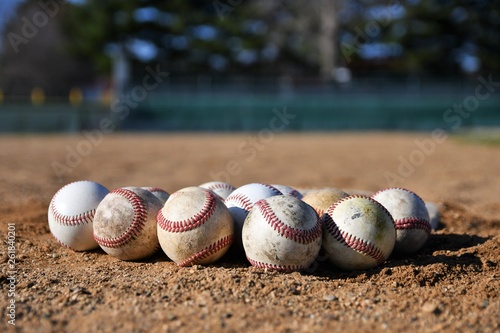 Close-up photo of a bunch of game used baseballs on a baseball infield on a sunny day with a baseball field in the background