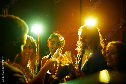 Group of hilarious young friends standing in circle and celebrating each other while drinking champagne at party in nightclub, disco lights effect