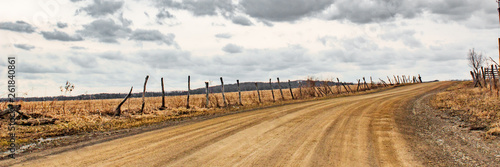 view up a brown dirt road with decaying barbed wire fence curving under cloudy gray sky