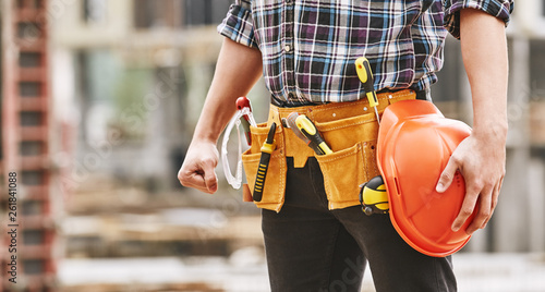 Safe work. Cropped photo of male professional builder with construction tools holding a safety red helmet while standing outdoor of construction site © Friends Stock