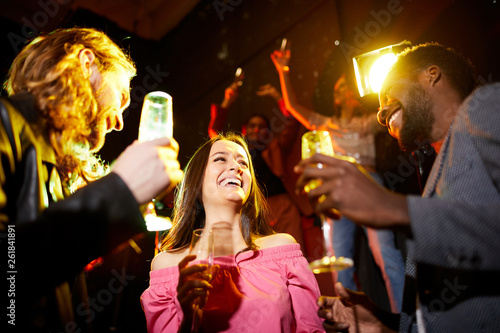Cheerful excited young lady in pink blouse drinking champagne and talking to two handsome guys at party