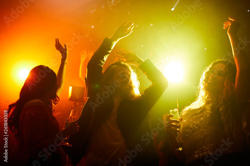 Positive excited youth in casual clothing drinking alcohol and dancing with raised arms in smoky dark dance hall