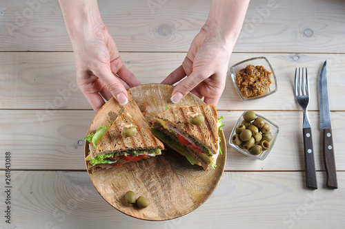 Two halves of a club sandwich on a wooden plate. The plate is held by two female hands. Olives, mustard and cutlery complement the composition. Light wooden background. View from above.