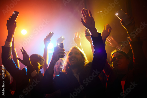 Fototapeta Naklejka Na Ścianę i Meble -  Happy excited young fans waving gadgets with flashlights turned on while greeting performer at concert in nightclub