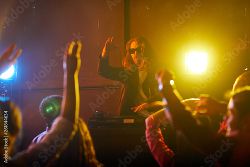 Handsome young bearded DJ performer using record player and gesturing hands while rocking crowd at party