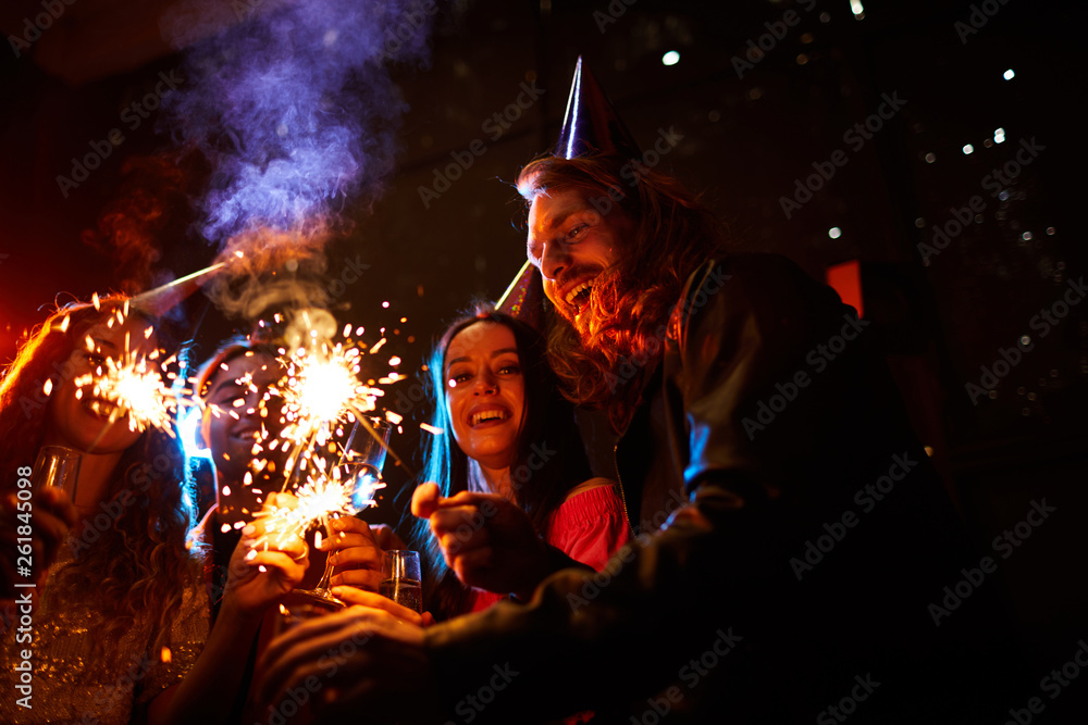 Happy excited young friends in party hats standing together in dark room and lighting sparklers while celebrating New Year Happy excited young friends in party hats standing together in dark room and lighting sparklers while celebrating New Year