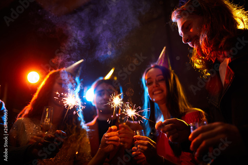 Close-up of positive young friends standing in dark room with colorful lights and lighting sparklers at birthday party