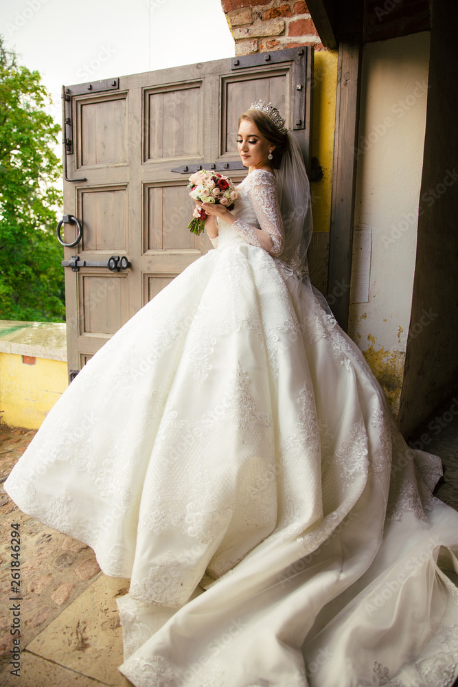 Fototapeta premium Beautiful Bride in a white Wedding Dress outdoors with a Bouquet of flowers and a veil
