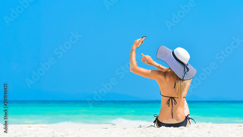 Cheerful young woman having fun taking smartphone selfie pictures of herself on tropical beach travel Caribbean holidays. Happy European girl model wearing fashion hat near sea. Summer lifestyle.