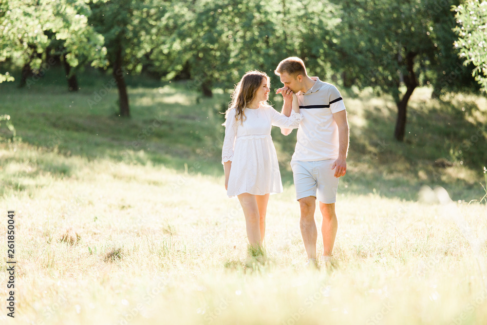 Fototapeta premium Young pretty couple in love walking in park. Handsome cheerful blonde girl in white dress hugging her boyfriend. Man and woman having fun outdoors