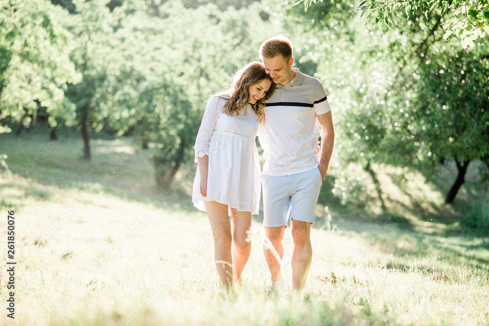 Fototapeta premium Young pretty couple in love walking in park. Handsome cheerful blonde girl in white dress hugging her boyfriend. Man and woman having fun outdoors