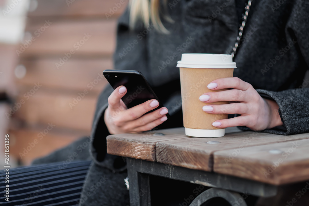 Close-up of woman's hands with a brown paper cup of coffee on an outdoor cafe bench. Uses a smartphone and the Internet. Close-up of woman's hands with a brown paper cup of coffee on an outdoor cafe bench. Uses a smartphone and the Internet.