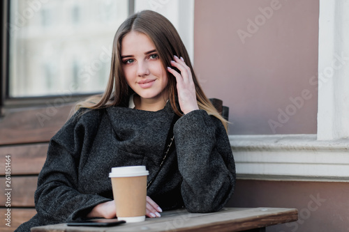 Young beautiful millenial girl in a coat is sitting on an outdor bench with a paper cup of coffee. Autumn or spring day, cool weather.