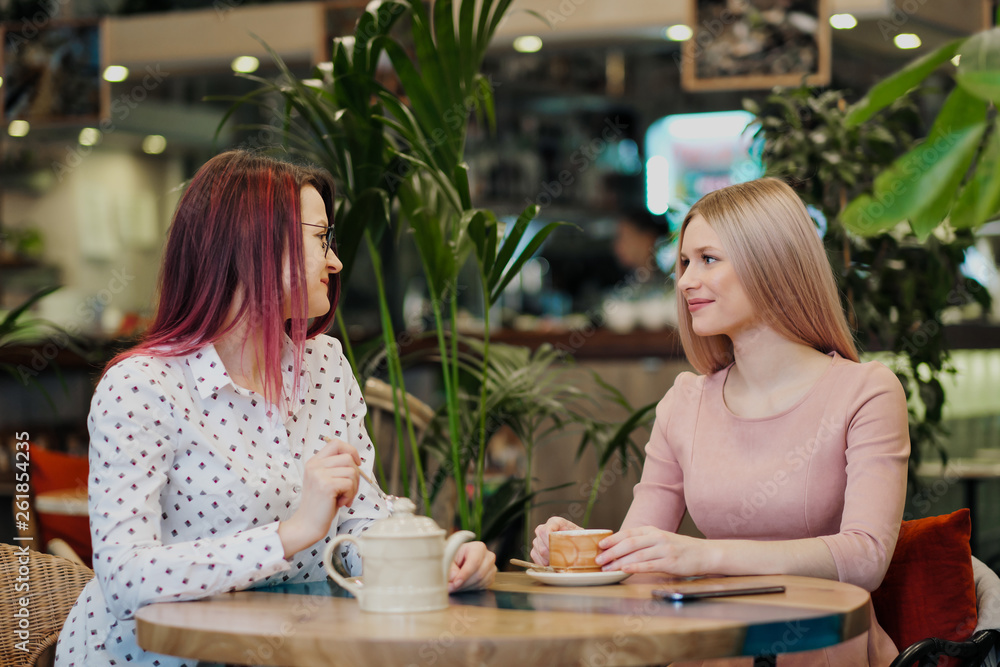 Two young beautiful girls sit in a green cafe at a table. Drink tea with croissants, chatting, laughing and taking pictures on a smartphone. Two young beautiful girls sit in a green cafe at a table. Drink tea with croissants, chatting, laughing and taking pictures on a smartphone.