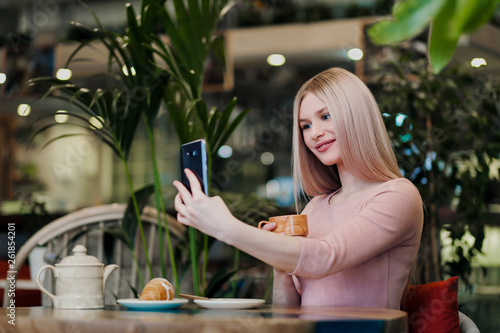 Young beautiful girl sitting in a green cafe at a table. Drinking tea with croissants, chatting, laughing and taking pictures on a smartphone.