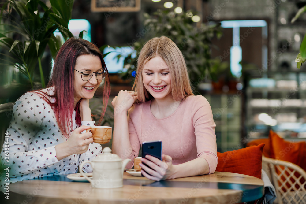 Two young beautiful girls sit in a green cafe at a table. Drink tea with croissants, chatting, laughing and taking pictures on a smartphone. Two young beautiful girls sit in a green cafe at a table. Drink tea with croissants, chatting, laughing and taking pictures on a smartphone.